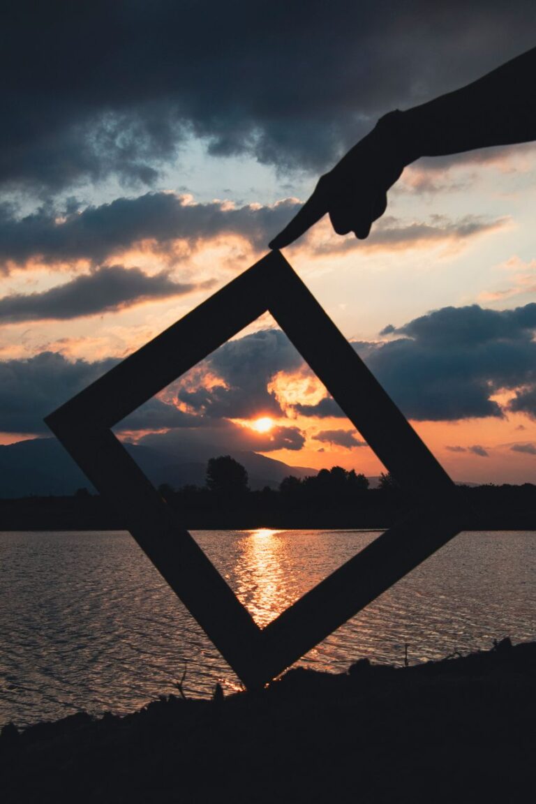 A hand frames the vibrant sunset over a serene lake, creating a scenic silhouette.